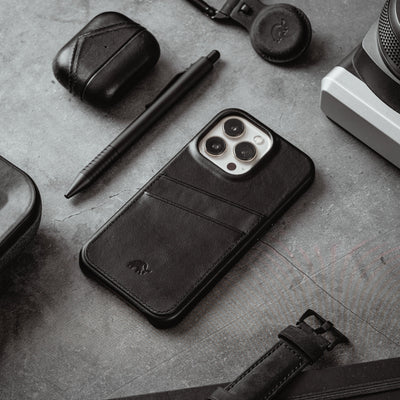 Black leather phone case with two card slots, styled with pen, earbuds case, camera and watch strap on textured surface.
