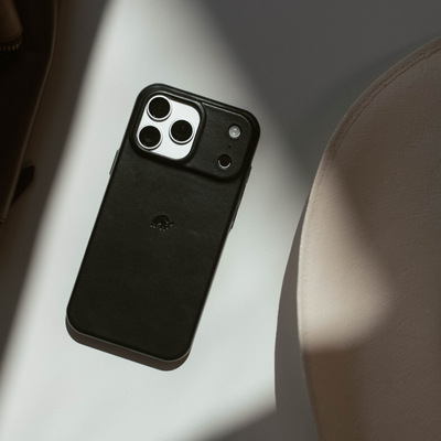 Black leather minimalist iPhone case with raised camera protector and small embossed logo on a pale tabletop.