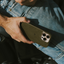 Hand holding iPhone with olive-green leather Maverick minimalist case over denim jeans, triple camera lenses visible